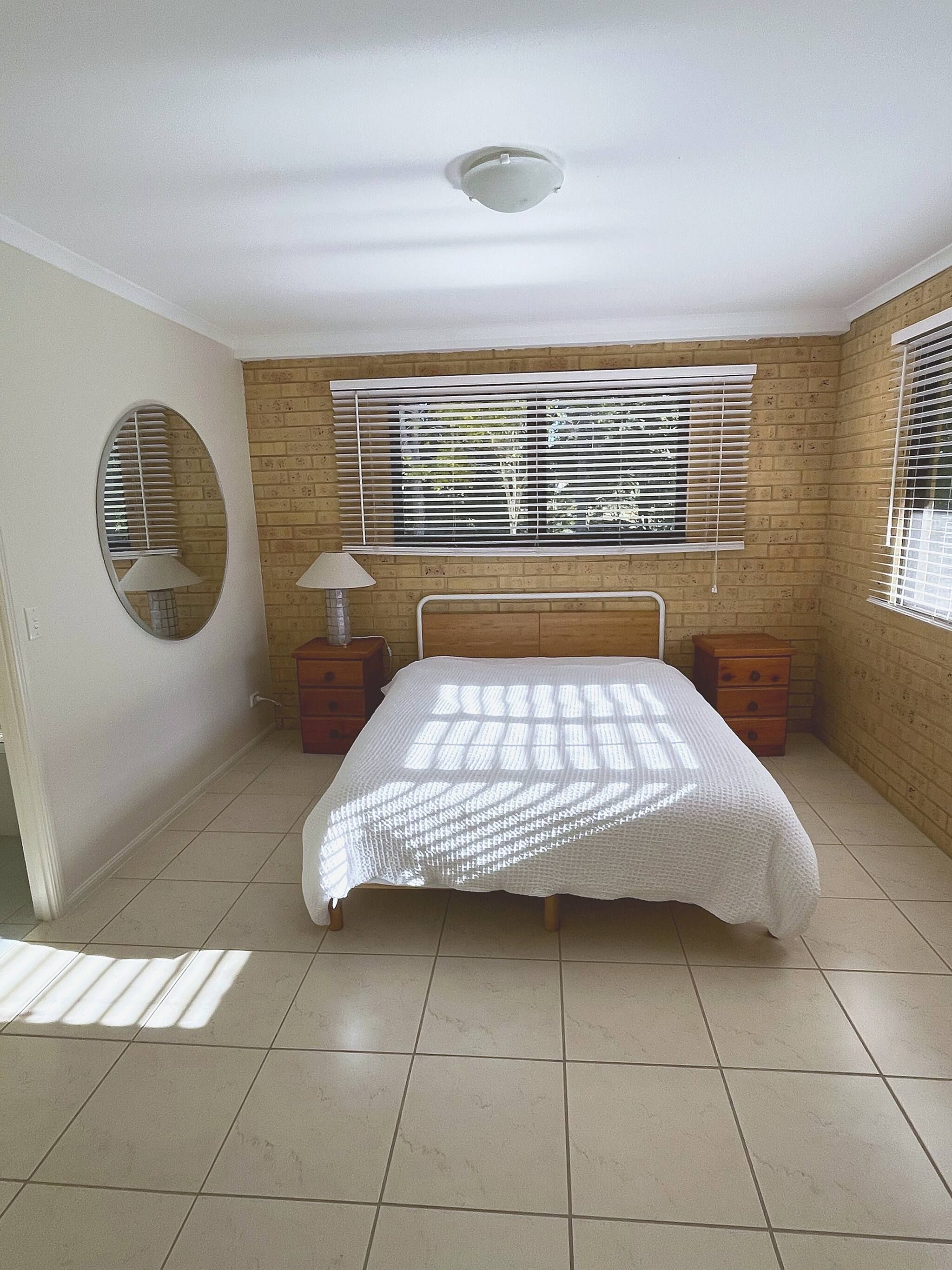 A bedroom with a bed, a window with blinds, a bedside lamp, and a circular mirror on the wall.