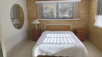 A bedroom with a bed, a window with blinds, a bedside lamp, and a circular mirror on the wall.