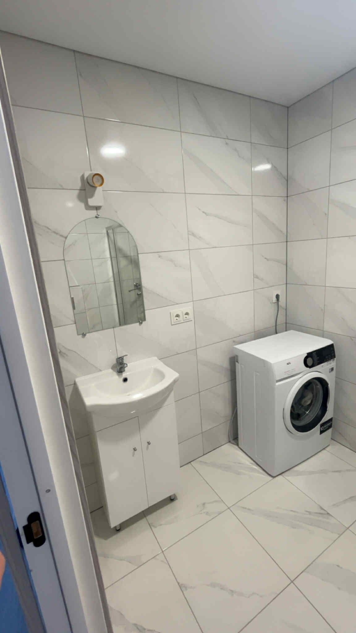 Un baño con un lavamanos blanco, espejo y lavadora pequeña.