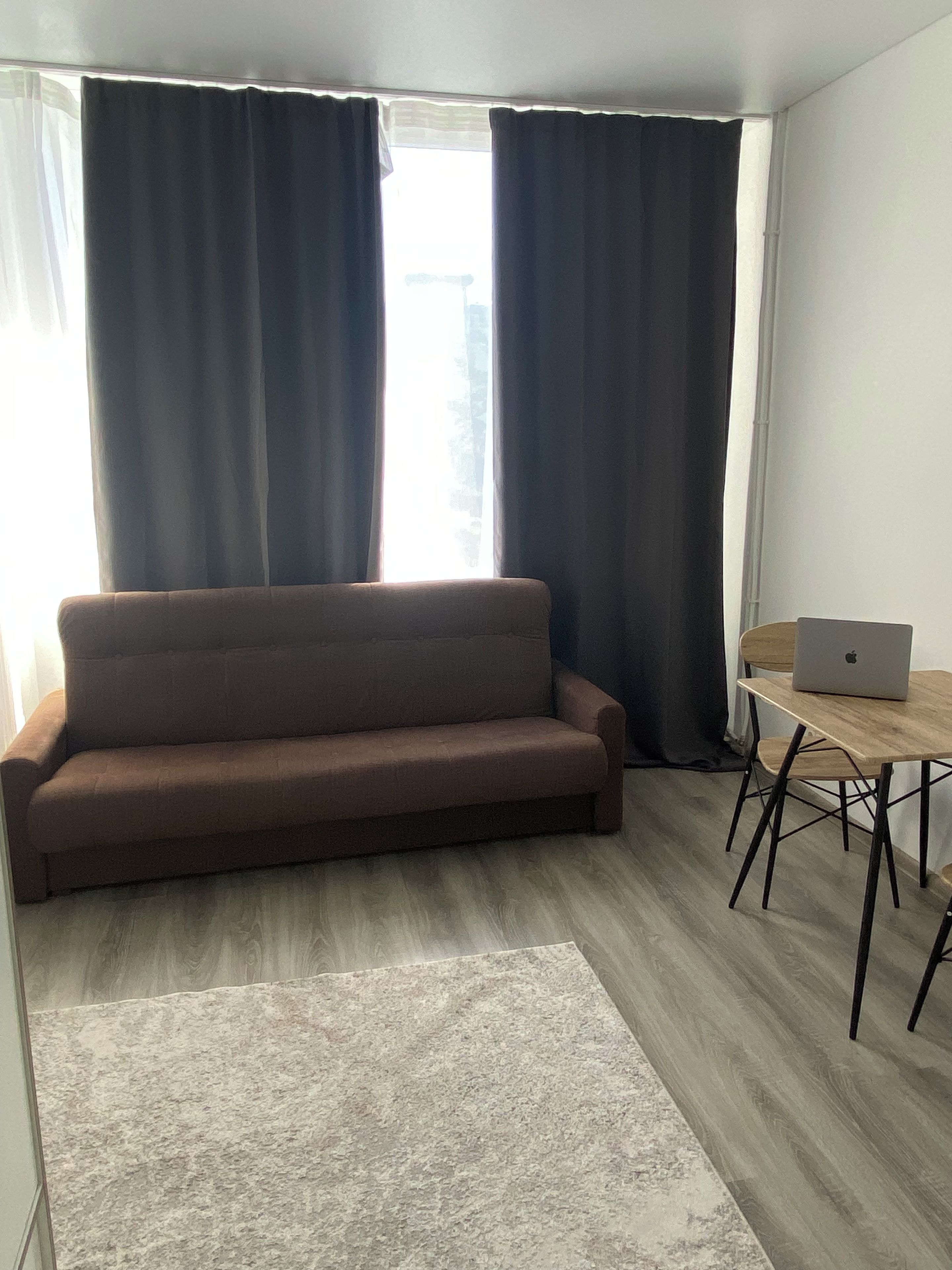Una sala de estar moderna con un sofá marrón, una pequeña mesa de madera con una computadora portátil, y un ventanal con cortinas.