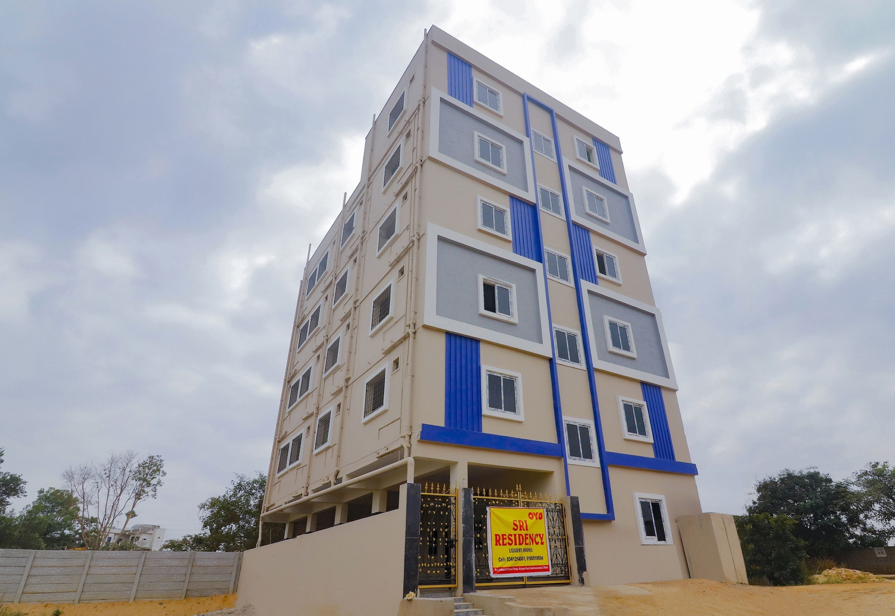 A multi-story residential building with a prominent blue and beige color scheme, featuring a gated entrance and a sign advertising Sri Residency.