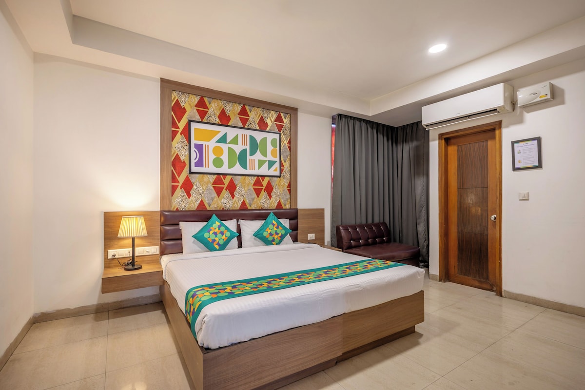 A hotel room with a bed, a nightstand, a lamp, a headboard with a colorful geometric pattern, a brown leather chair, a wooden door, and an air conditioning unit.