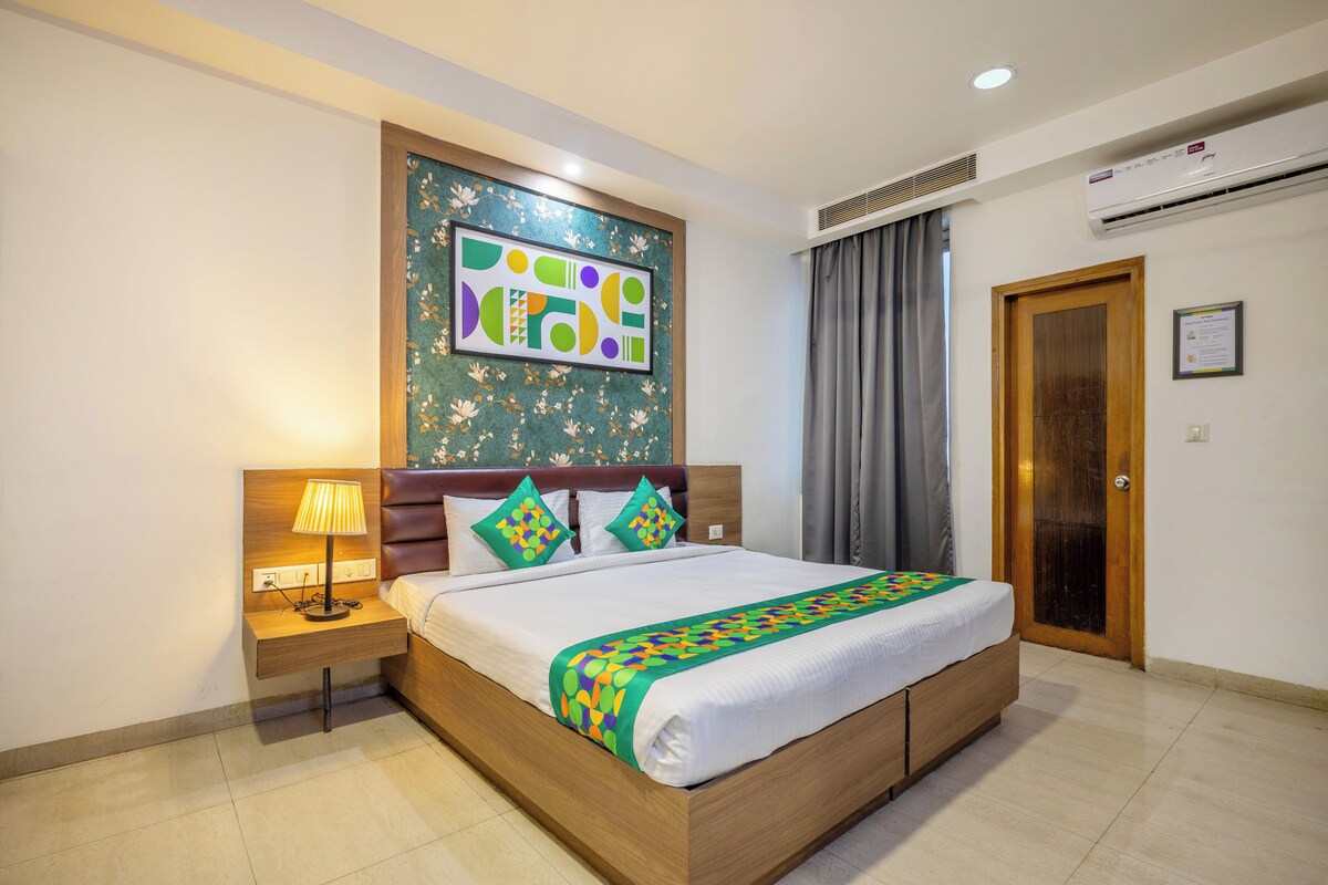 A hotel room with a wooden bed, a nightstand with a lamp, a wall-mounted air conditioner, and a framed abstract artwork.