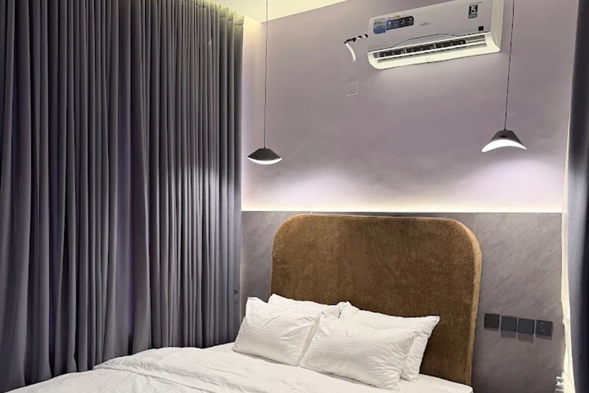 A modern bedroom with a large bed, a bedside table, an air conditioning unit, and a curtain.