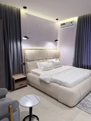 A modern bedroom with a large bed, bedside tables, a small round table, and a ceiling-mounted air conditioning unit. - Liam's Signature Apartment (Lagos)