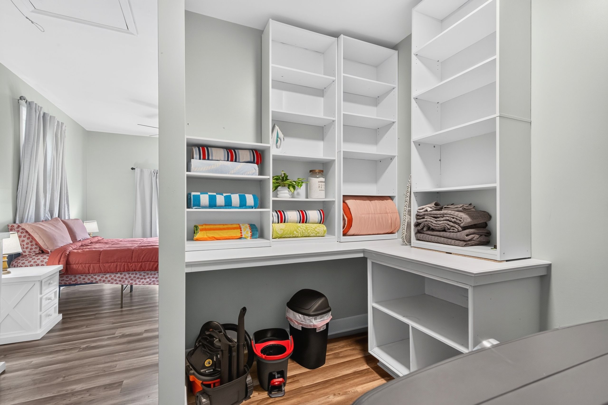 A bedroom with a bed, a nightstand, a desk, and a shelving unit with folded towels and a trash bin.