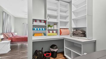 A bedroom with a bed, a nightstand, a desk, and a shelving unit with folded towels and a trash bin.