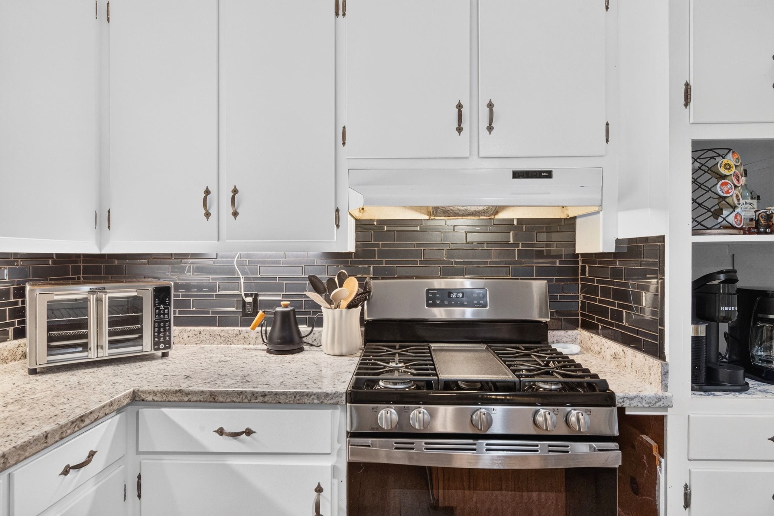 A kitchen with white cabinets, a black tiled backsplash, a stainless steel stove, and a coffee maker.