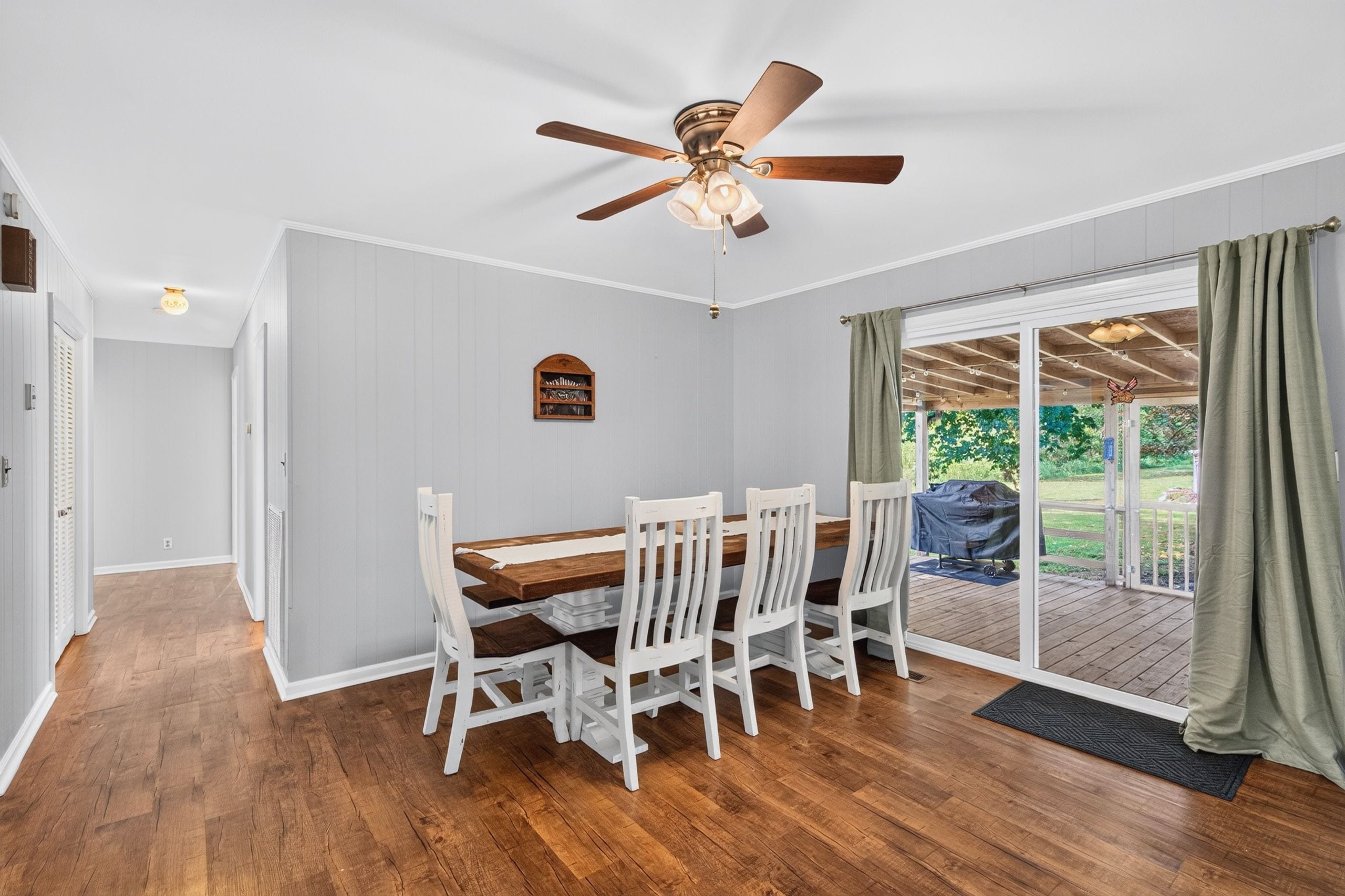 A dining area with a wooden table and white chairs, a ceiling fan, and a view through a glass door to an outdoor patio.