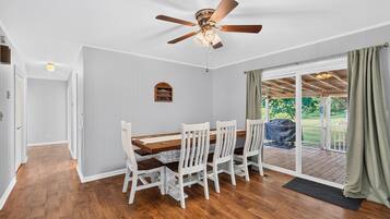 A dining area with a wooden table and white chairs, a ceiling fan, and a view through a glass door to an outdoor patio.