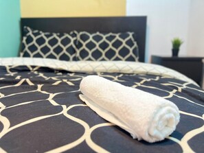 Een opgemaakt bed met een opgerolde witte handdoek, een bed met een dessin en een hoofdbord met een geometrisch patroon.