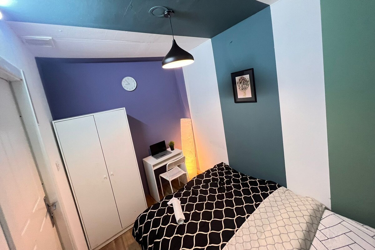 Een slaapkamer met een bed, bureau, stoel, kledingkast, klok en een aan de muur hangend schilderij.