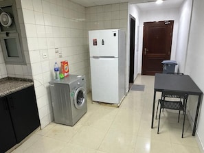 A compact kitchen with a washing machine, a refrigerator, and a dining table with a chair. - Double Bed Partition Room in Al Barsha1 (Dubai)