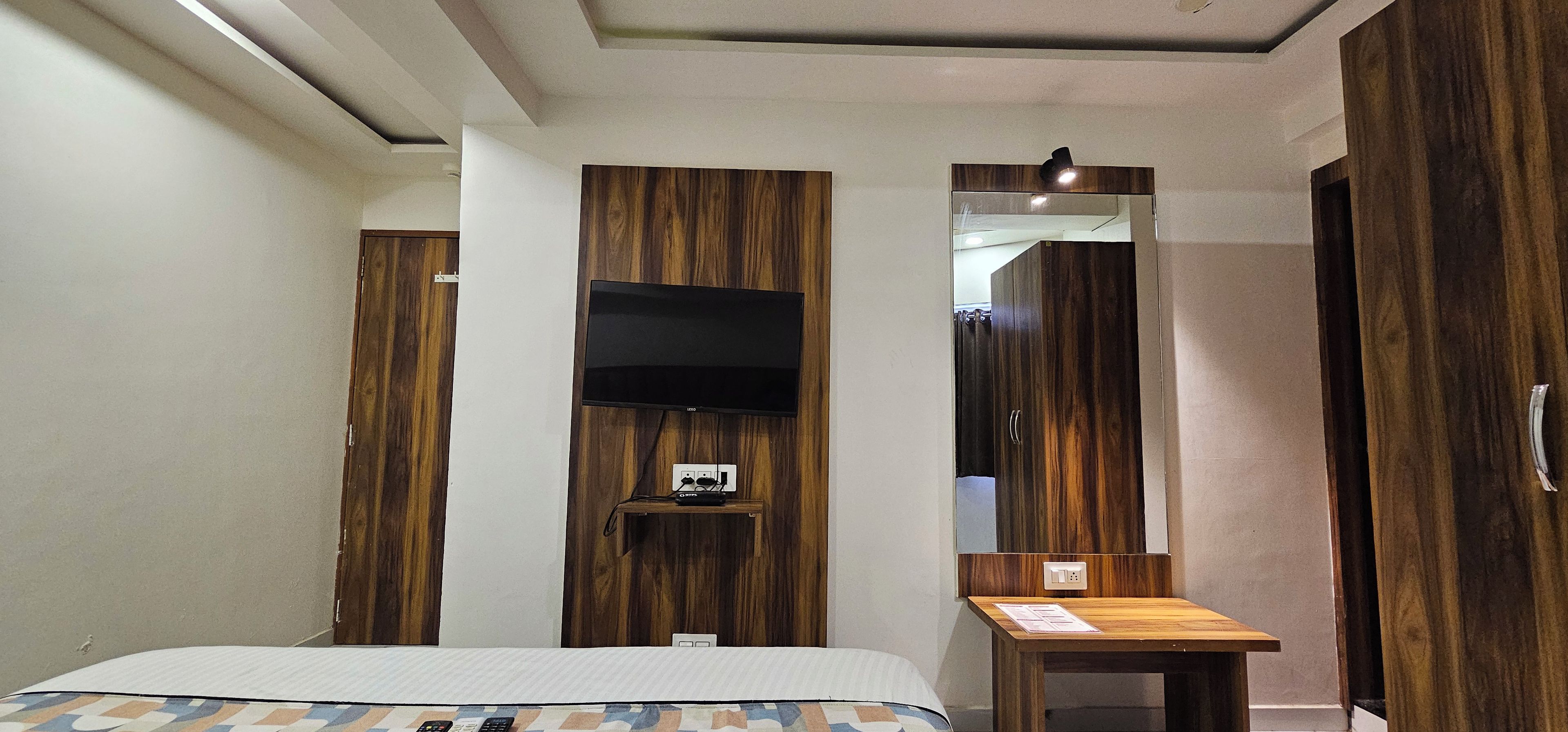 A hotel room with a bed, a desk, a mirror, and a television mounted on a wooden panel wall.