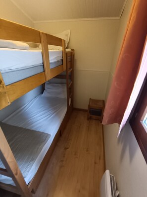 Habitación pequeña con dos literas, una mesita de noche de madera, un calentador y una ventana con cortinas.