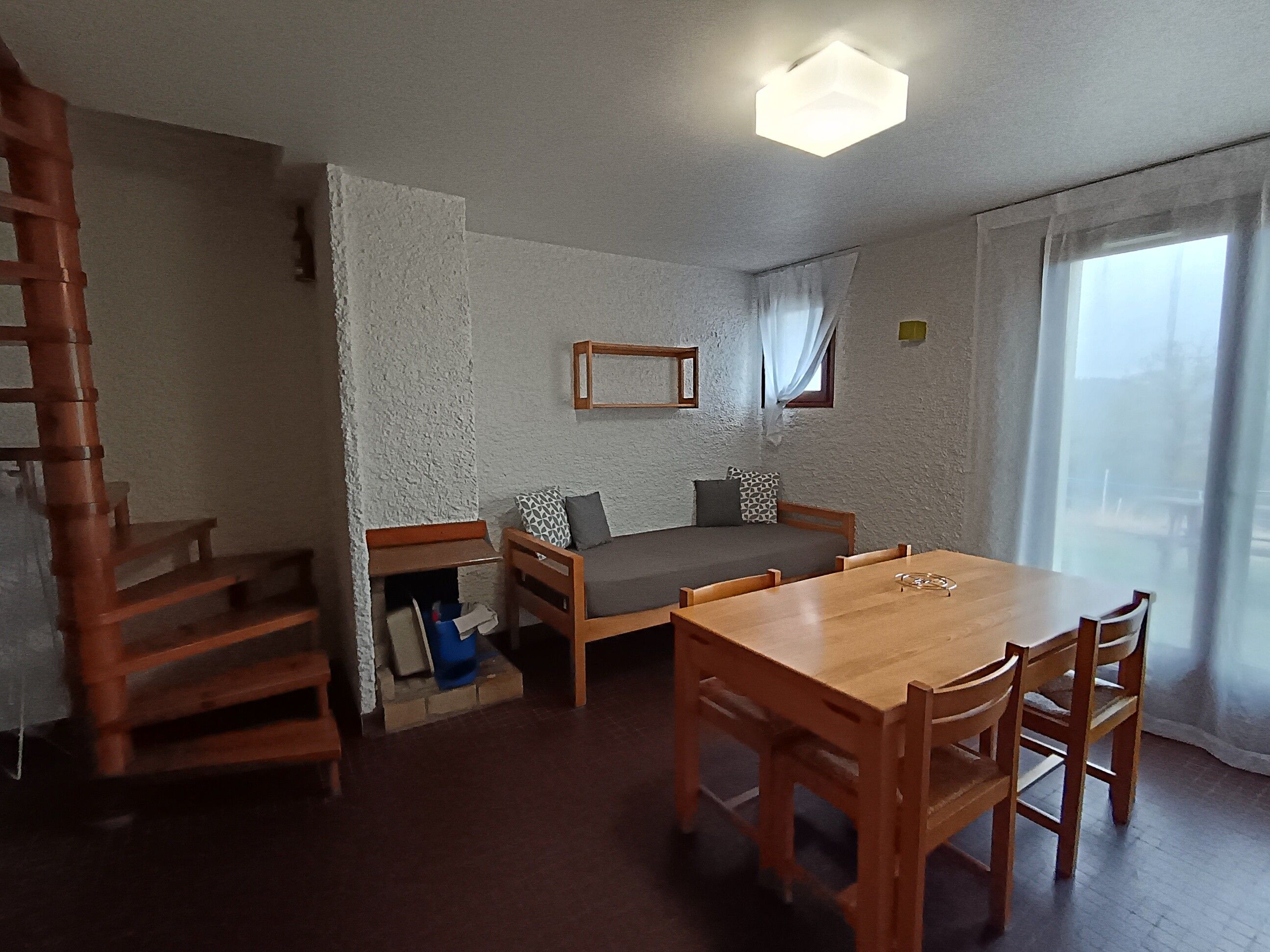 A room with a wooden dining table, a sofa, a staircase, and a window with curtains.