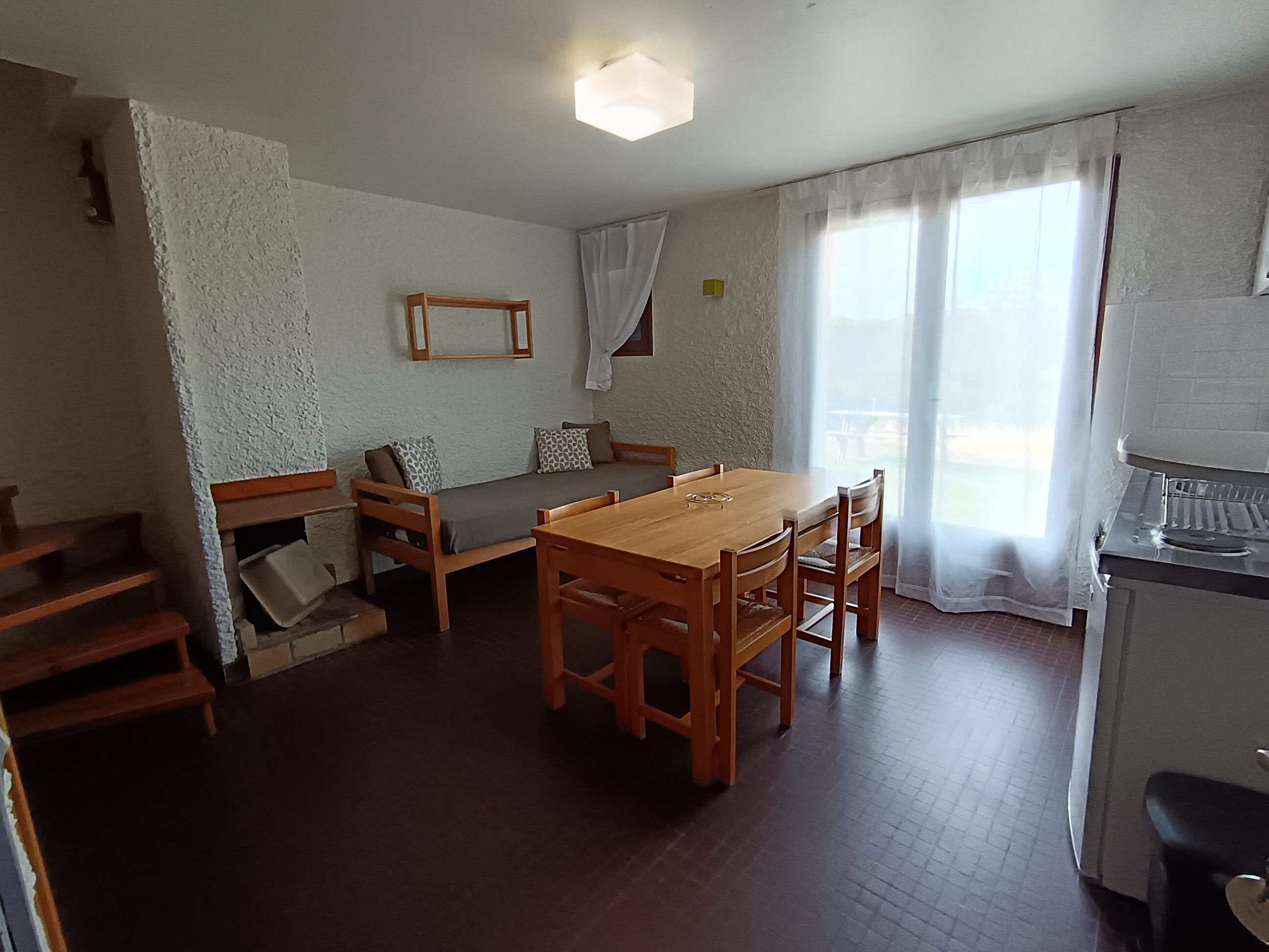 A room with a wooden dining table, a sofa with patterned cushions, a small kitchen area, and a staircase.