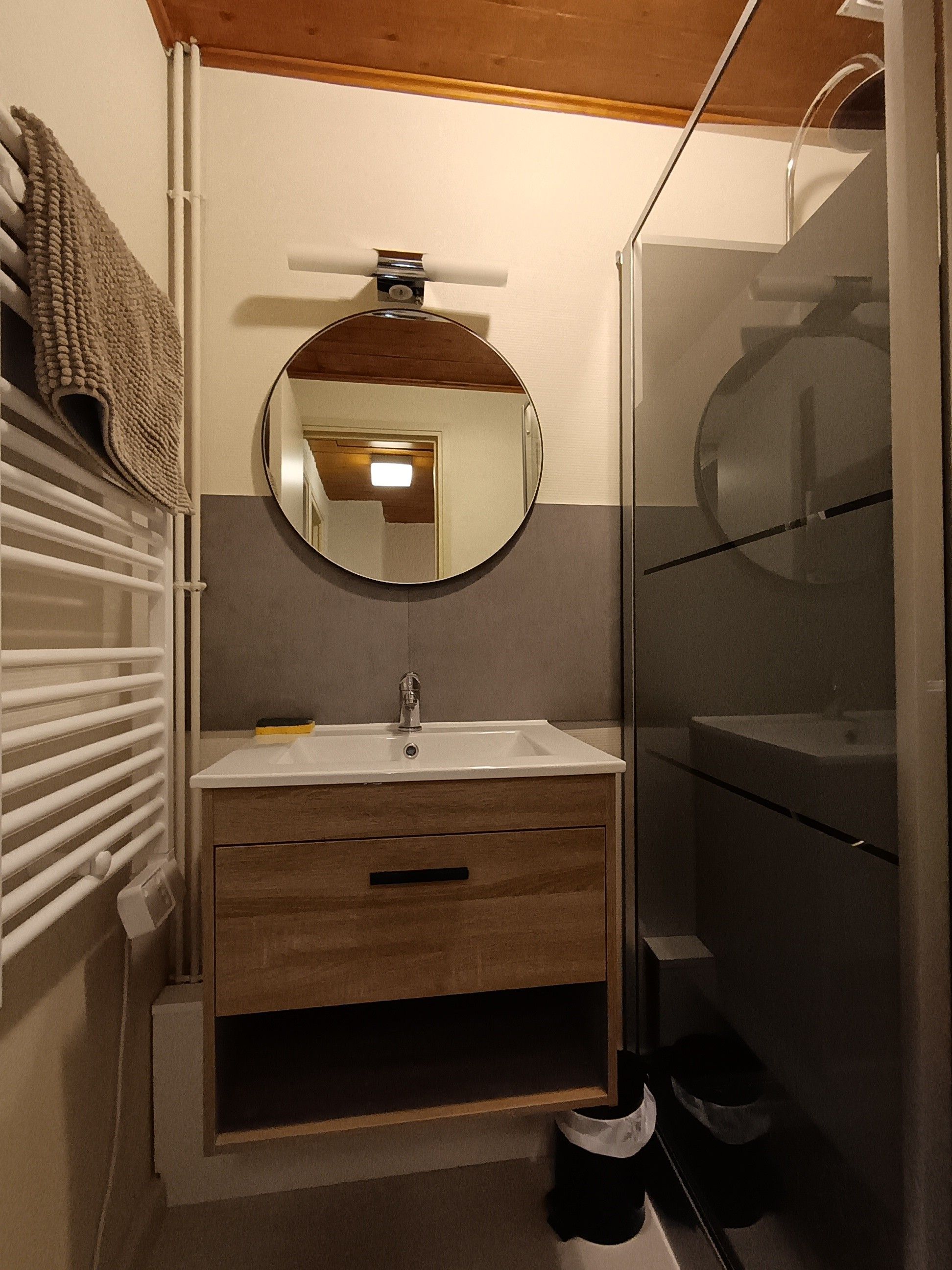 Une salle de bain avec un miroir rond, un lavabo, un meuble en bois et une cabine de douche en verre.