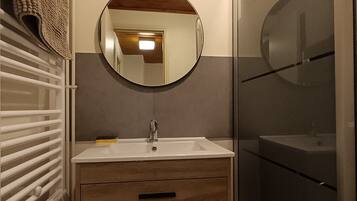 Une salle de bain avec un miroir rond, un lavabo, un meuble en bois et une cabine de douche en verre.