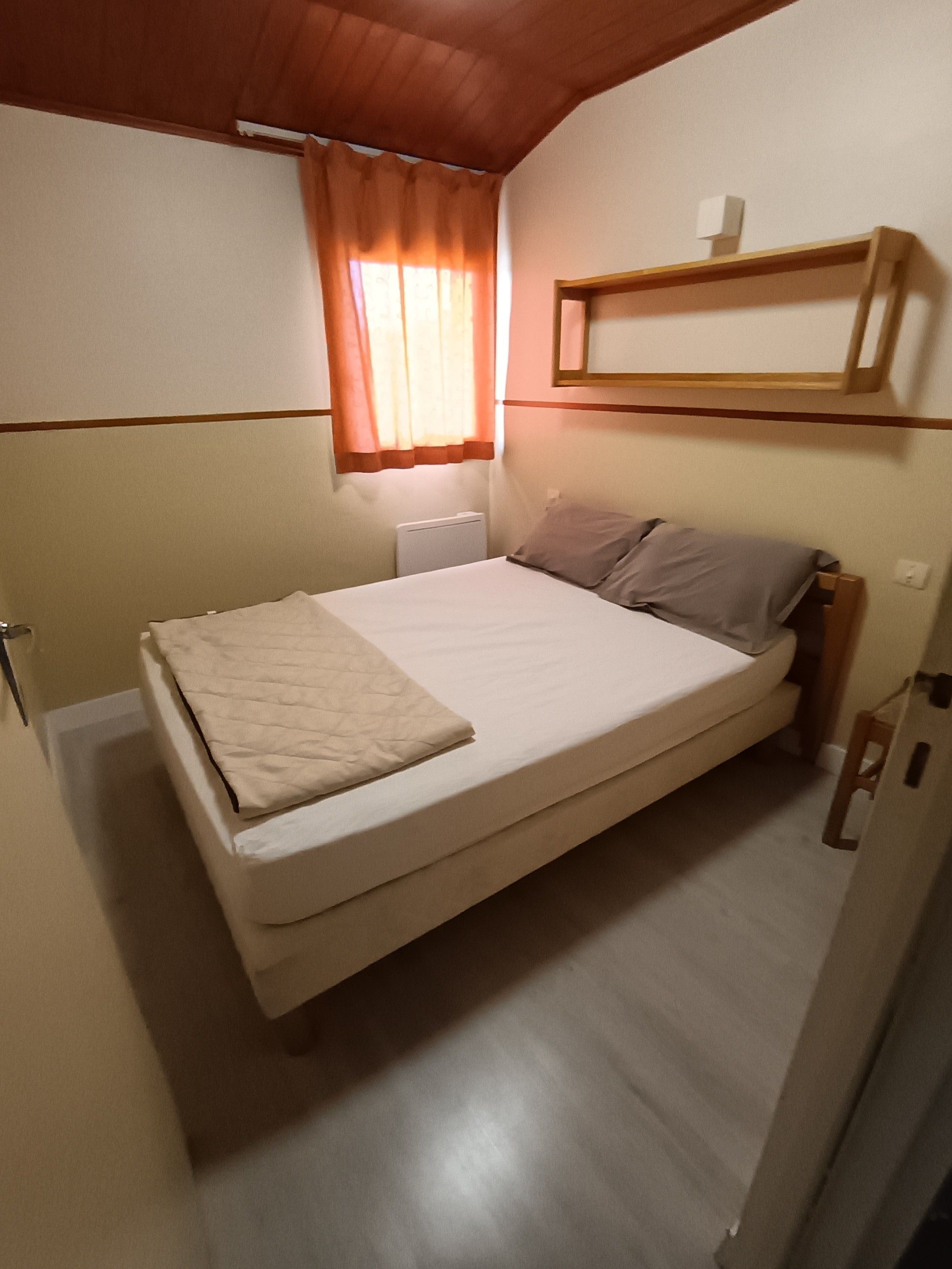 Ein Einzelbett mit einer gesteppten Decke, ein Heizkörper, ein Fenster mit Vorhängen und ein Holzregal über dem Bett.