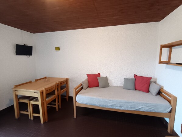 Una habitación pequeña y acogedora con una mesa y sillas de madera, una cama individual con almohadas y un televisor montado en la pared.