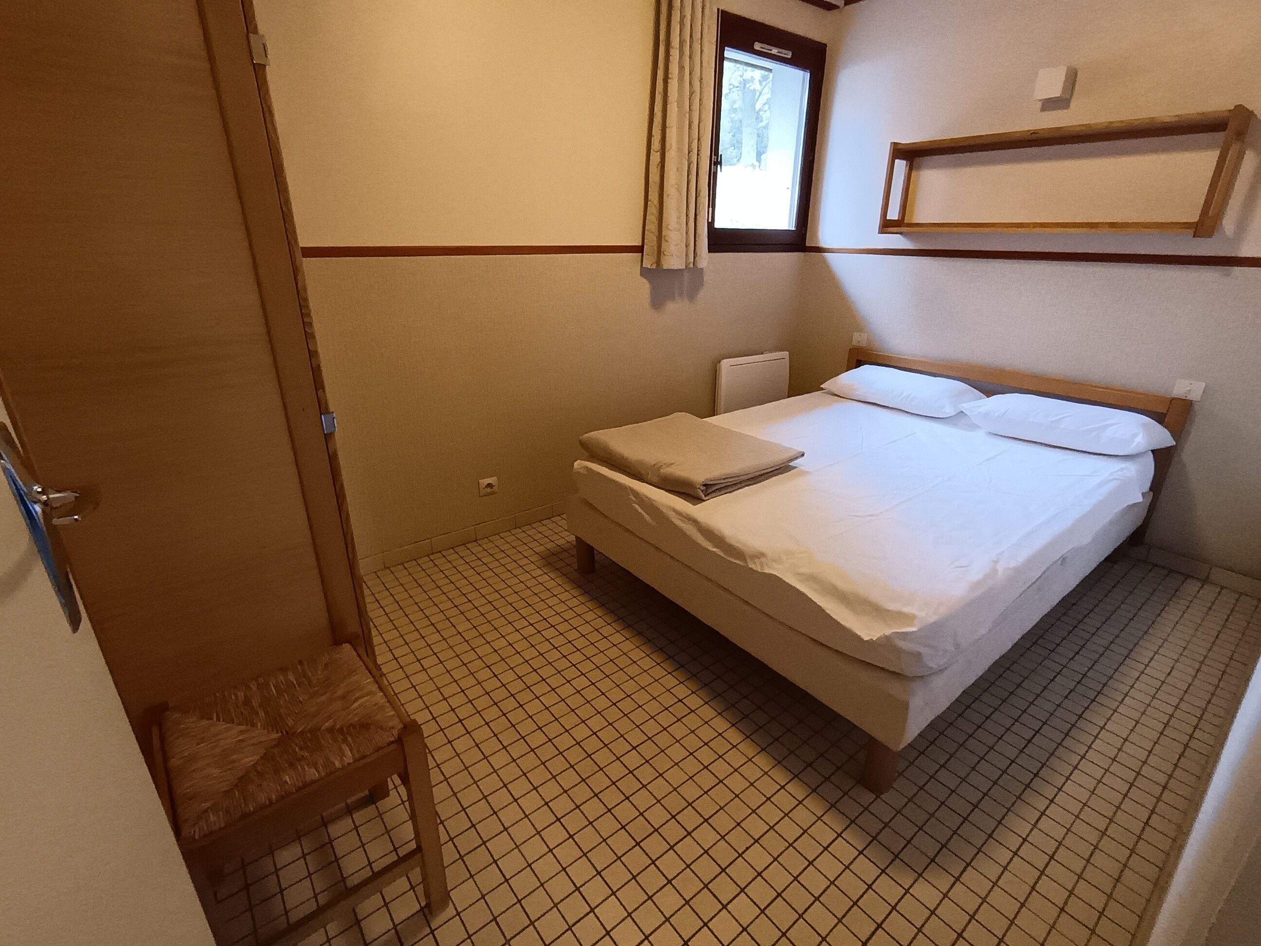 A small, single-bed room with a wooden headboard, a bedside table, and a window with curtains.