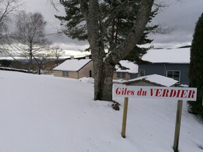 Un cartel con el texto 'Gîtes du VERDIER' en un entorno rural, con suelo cubierto de nieve y casas al fondo.
