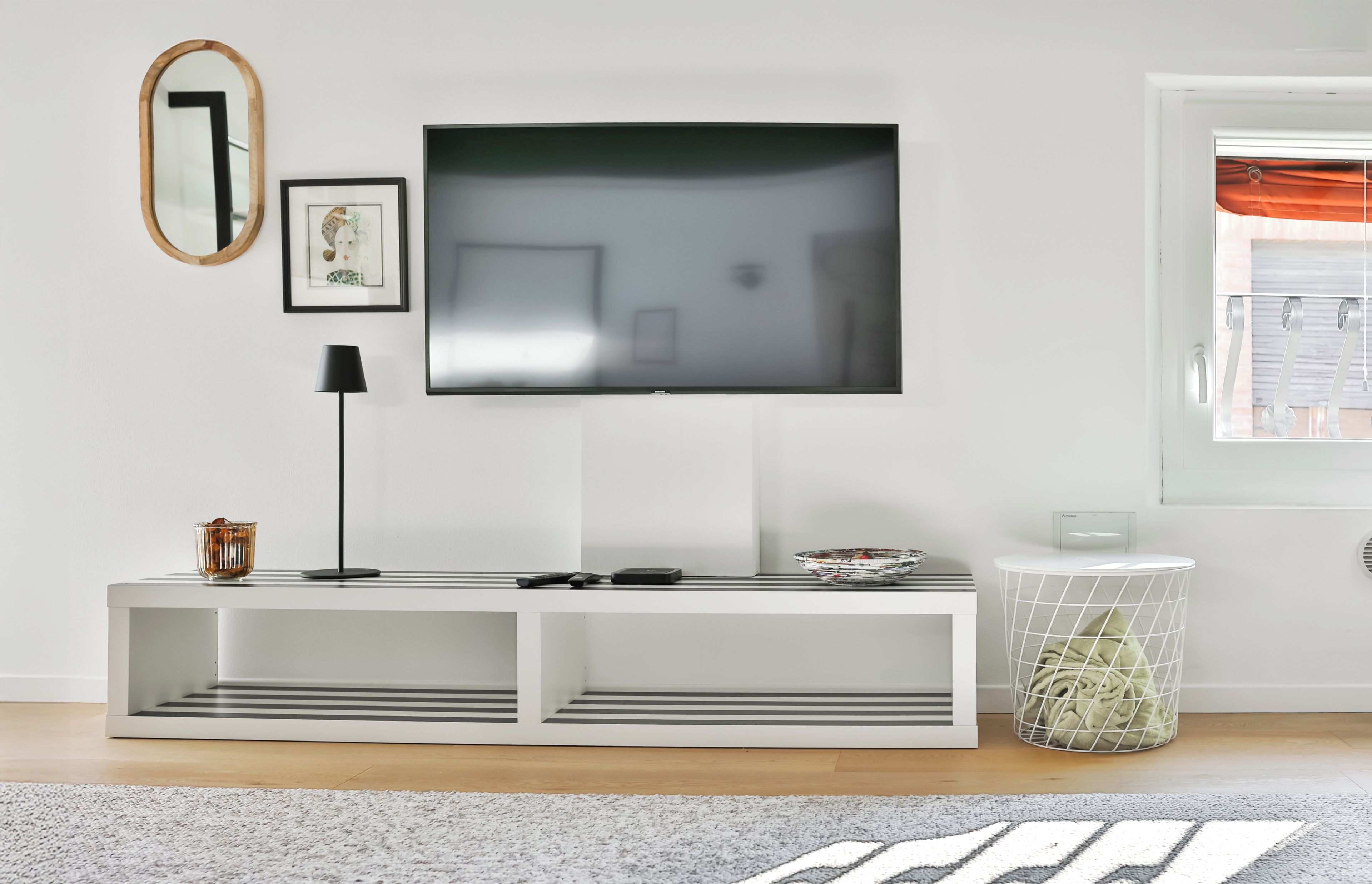 Ein modernes Wohnzimmer mit einem wandmontierten Flachbildfernseher, einem gerahmten Bild, einer Stehlampe und einer weißen Unterhaltungsanlage mit einer Glasvase und Fernbedienungen.