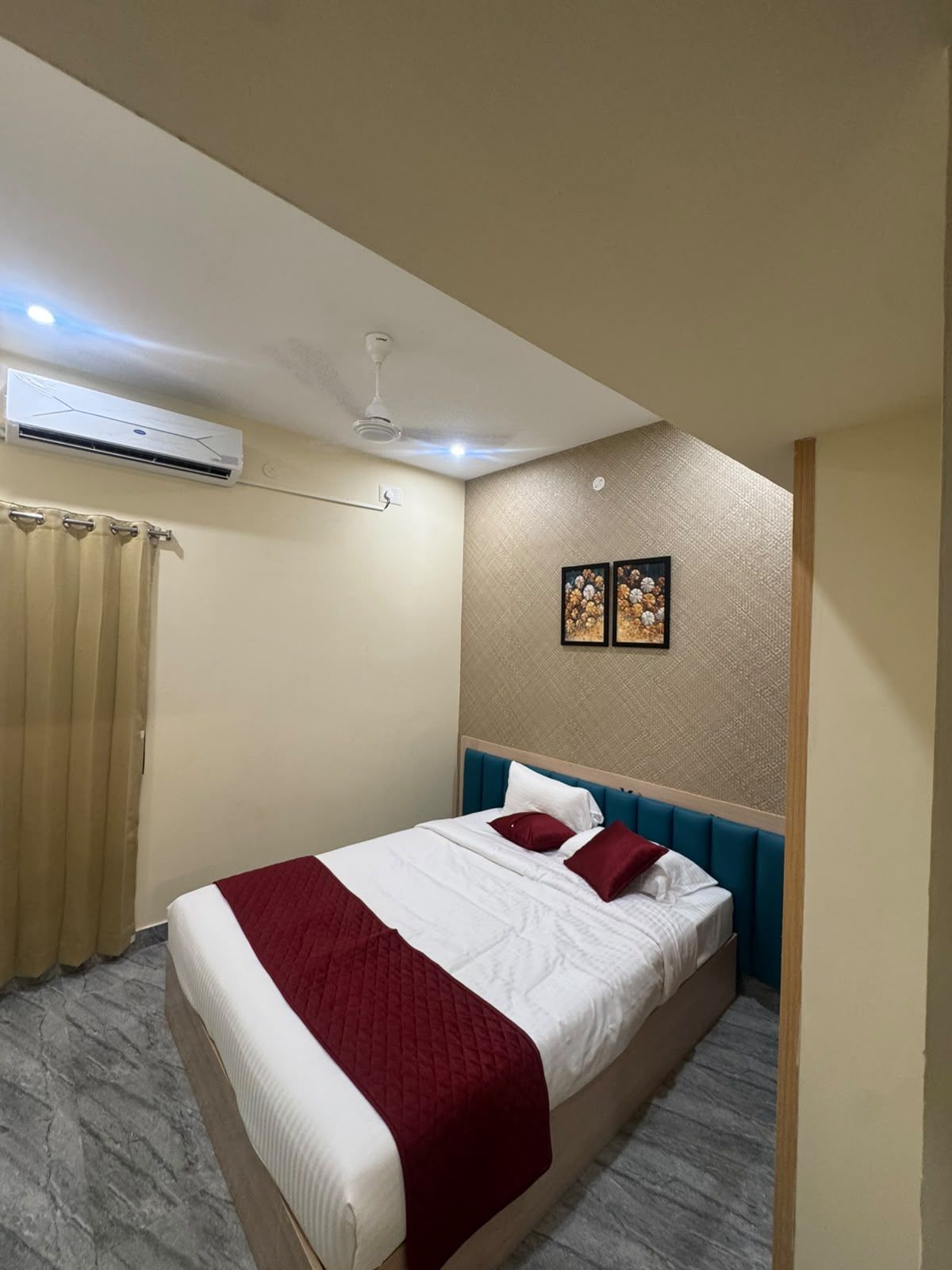 A hotel room with a bed, a ceiling fan, an air conditioning unit, a window with curtains, and two wall-mounted pictures.