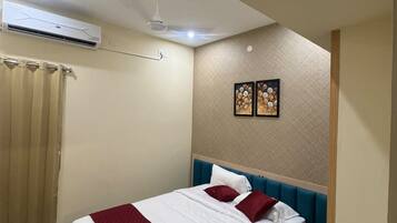 A hotel room with a bed, a ceiling fan, an air conditioning unit, a window with curtains, and two wall-mounted pictures.