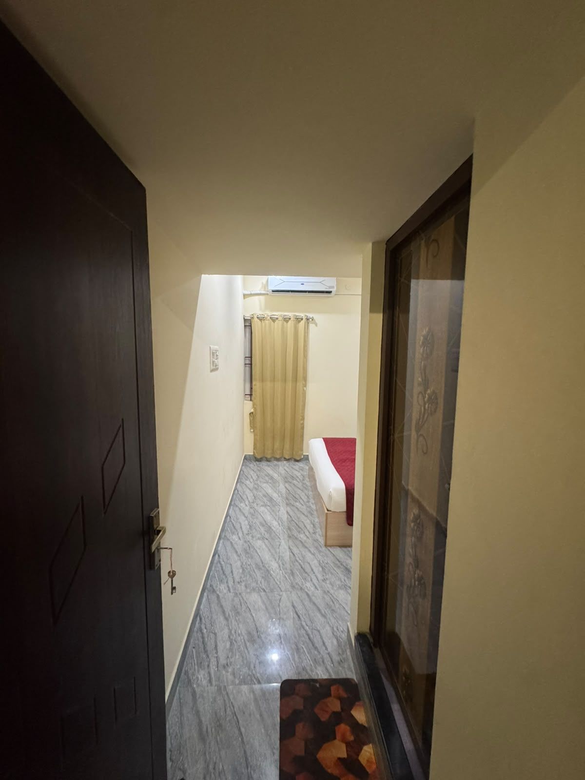 Un pasillo estrecho con una puerta a la izquierda, una cama con una colcha roja en el centro y un tapete estampado al final.