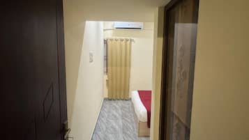 Un pasillo estrecho con una puerta a la izquierda, una cama con una colcha roja en el centro y un tapete estampado al final.