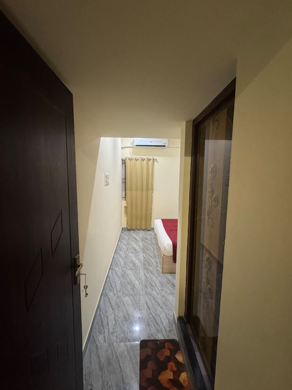 Un pasillo estrecho con una puerta a la izquierda, una cama con una colcha roja en el centro y un tapete estampado al final.