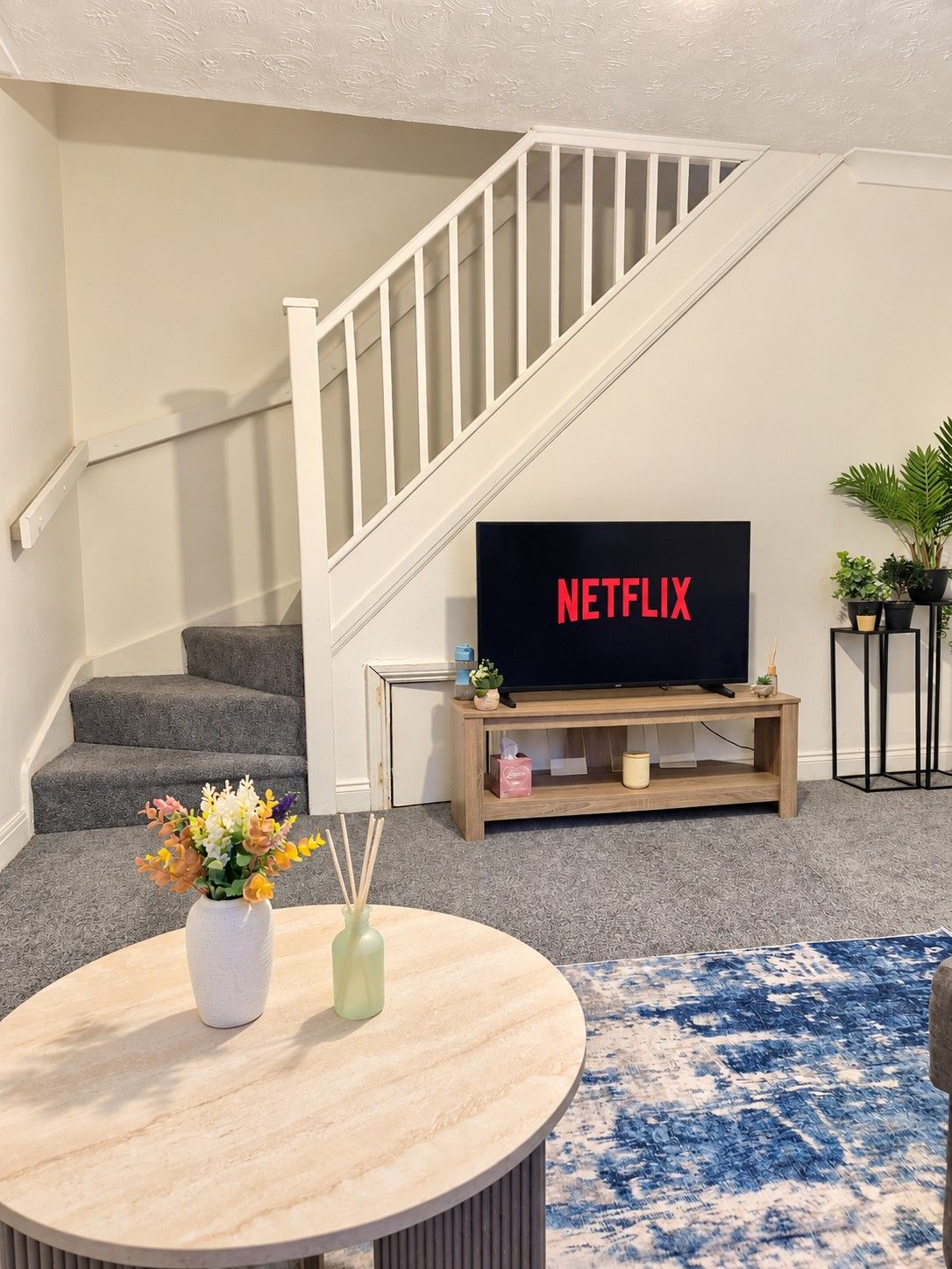 Ein Wohnzimmer mit Treppe, einem Flachbildfernseher auf einem Holzbodenständer, einem runden Tisch mit Blumenvase und einem blauen Teppich mit Muster.