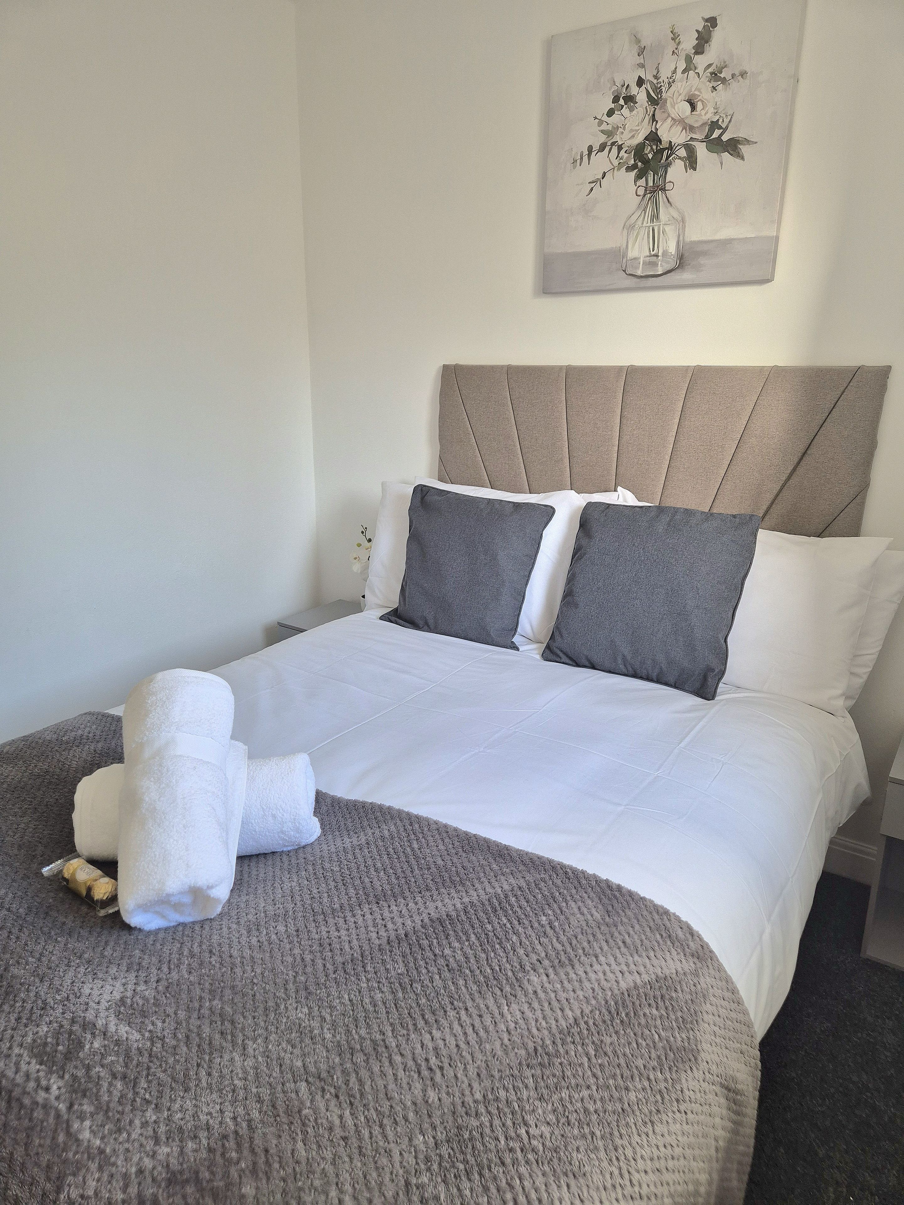 A neatly made bed with white and gray bedding, a gray throw blanket, and two rolled towels.