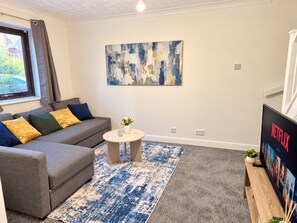 A living room with a grey sofa, a coffee table, a blue and white rug, a wall art piece, and a Netflix poster. - Cosy 2 Bedroom Home in Great Location (Shenley Lodge)