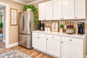 A modern kitchen with a stainless steel refrigerator, white cabinetry, a coffee station, and a framed inspirational quote on the wall. - Larry's Love - A Coastal Retreat Near the Beach (Wilmington)
