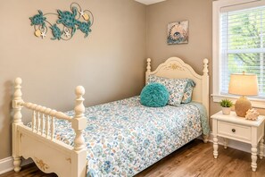 A bedroom with a floral bedspread, a round teal pillow, a nightstand with a lamp, and a wall art piece. - Larry's Love - A Coastal Retreat Near the Beach (Wilmington)