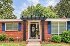 A two-story house with a brick facade, a black porch with a welcome mat, and a small garden area. - Larry's Love - A Coastal Retreat Near the Beach (Wilmington)
