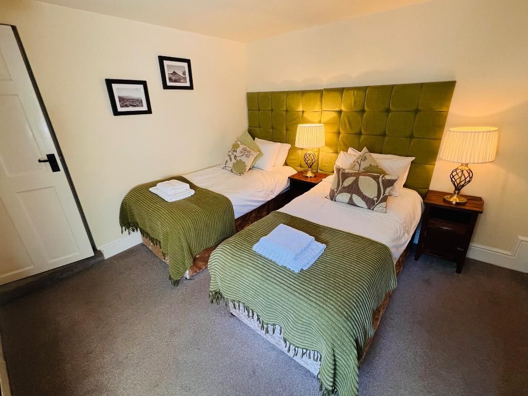 A hotel room with two beds, each with a green bedspread and white linens. There are two bedside tables with lamps, and the room has a neutral color scheme.