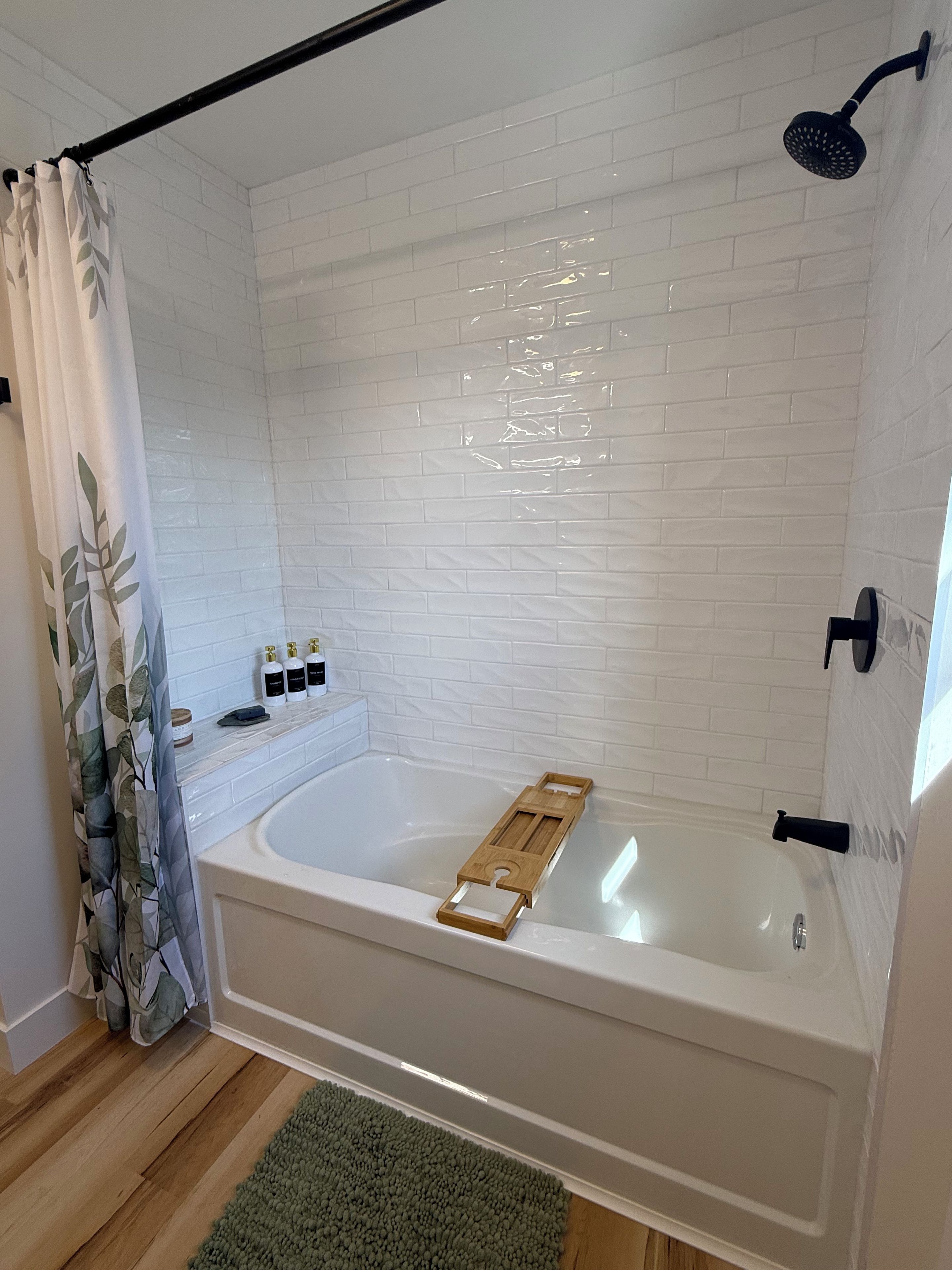 Un baño moderno con ducha de azulejos blancos, bañera con bandeja de madera y alfombra verde en el suelo.