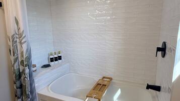 Un baño moderno con ducha de azulejos blancos, bañera con bandeja de madera y alfombra verde en el suelo.