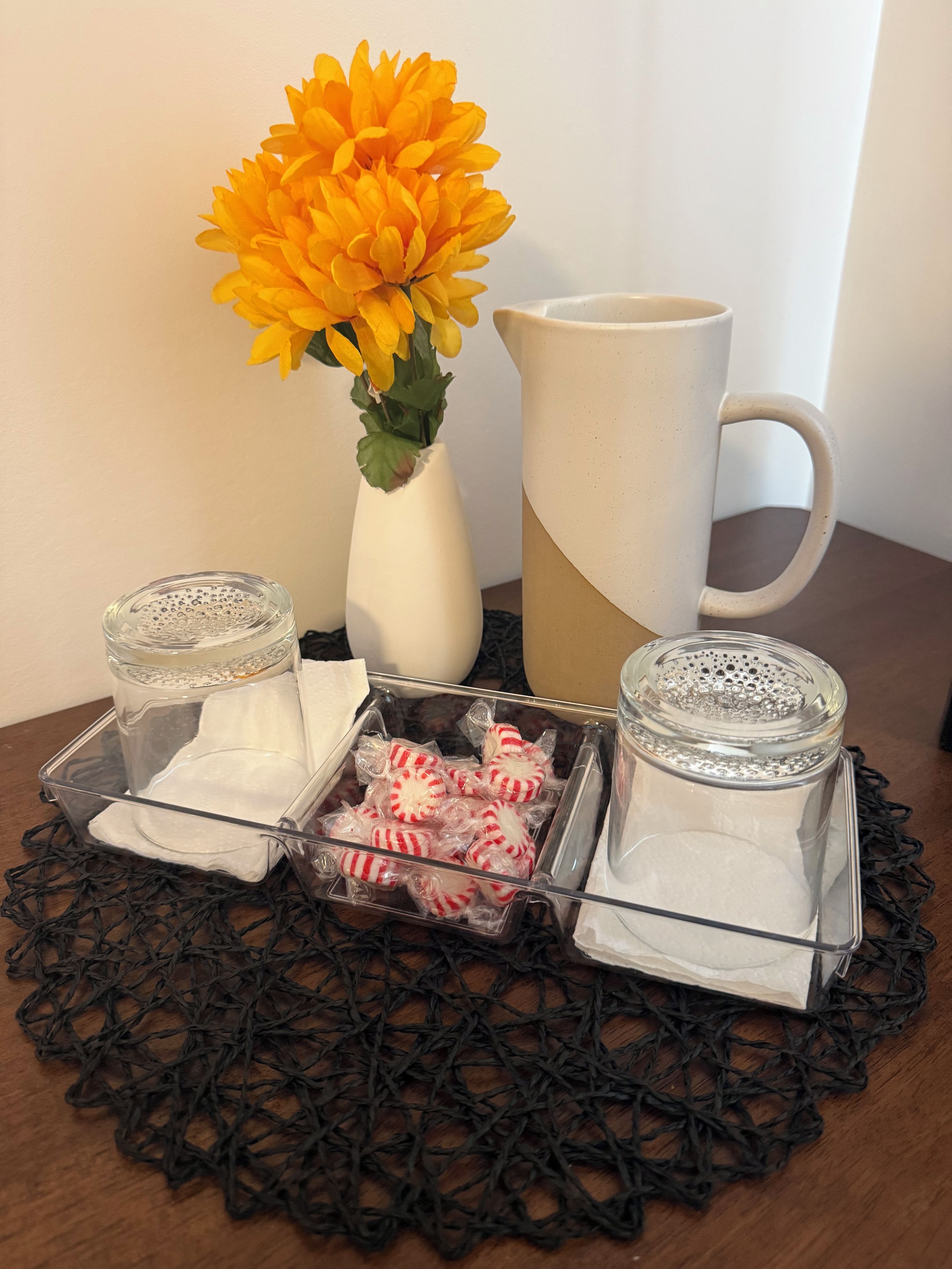 Et bord med et klart glasstage, en hvid vase med gule blomster, en hvid kop og et fad med slik indpakket i plastic.