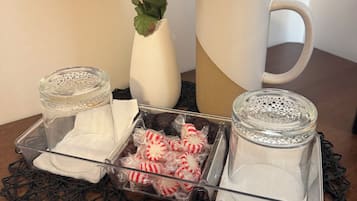 A table with a clear glass candle holder, a white vase with yellow flowers, a white mug, and a tray with candy wrapped in plastic.