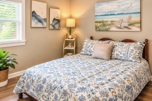 Un dormitorio con cama, mesita de noche, lámpara, ventana con persianas y una pared decorada con una obra de arte.
