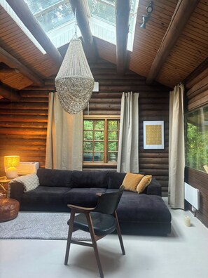 A log cabin interior with a dark-colored sofa, a hanging chandelier, and a window with curtains. - Timber-framed house with sedum roof in nature (Frederiksværk)