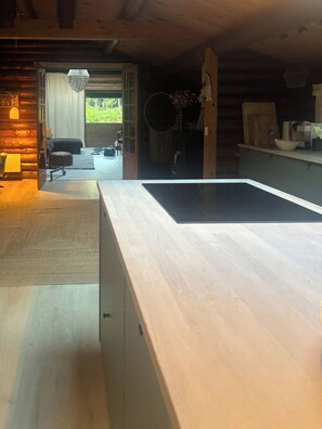 A modern kitchen with a large countertop, wooden cabinets, and a view into a living area with a sofa and a dining table. - Timber-framed house with sedum roof in nature (Frederiksværk)