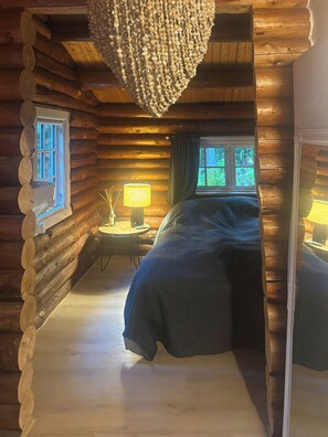 Quarto de cabana de tronco com uma cama, mesa de cabeceira, luminária e janela com cortinas.