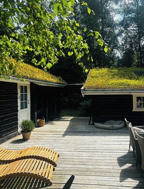 A wooden deck with a covered seating area, a small garden with yellow flowers, and a building with a green roof. - Timber-framed house with sedum roof in nature (Frederiksværk)