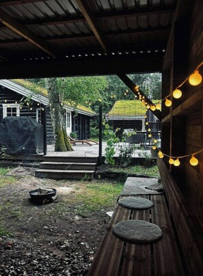 A covered outdoor area with wooden benches, string lights, and a view of a house with a green roof. - Timber-framed house with sedum roof in nature (Frederiksværk)